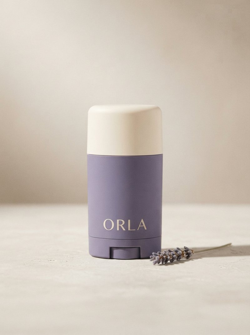 A lavender deodorant stick with a cream cap standing upright on a warm oat plaster surface beside a dried lavender sprig