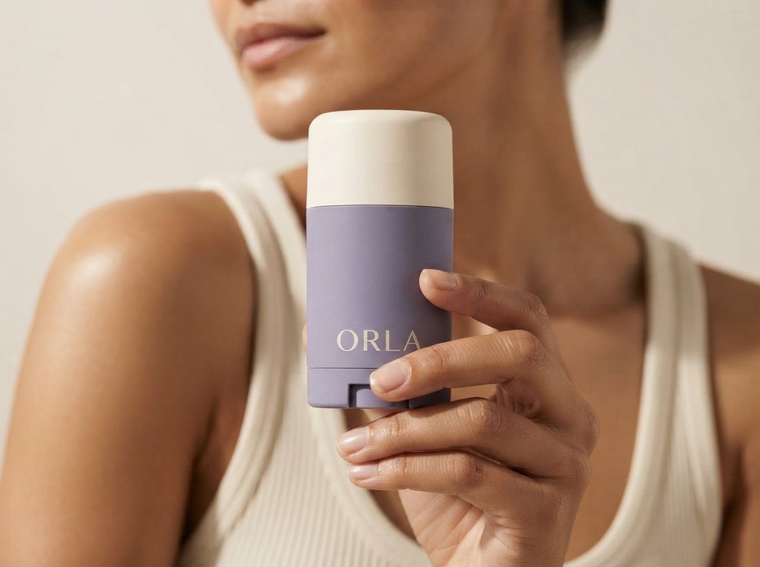 Close up of a woman holding the lavender deodorant stick at shoulder height against a warm cream background, the product filling most of the frame