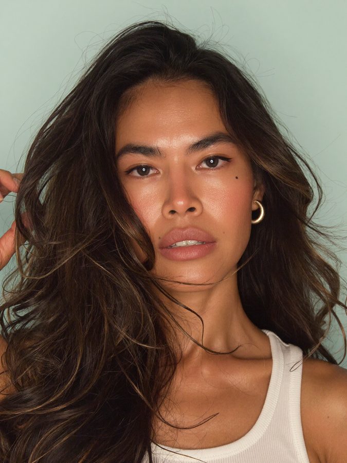 The AI brand model in a white ribbed tank against a pale seafoam backdrop, hair windswept, hand raised to her hair