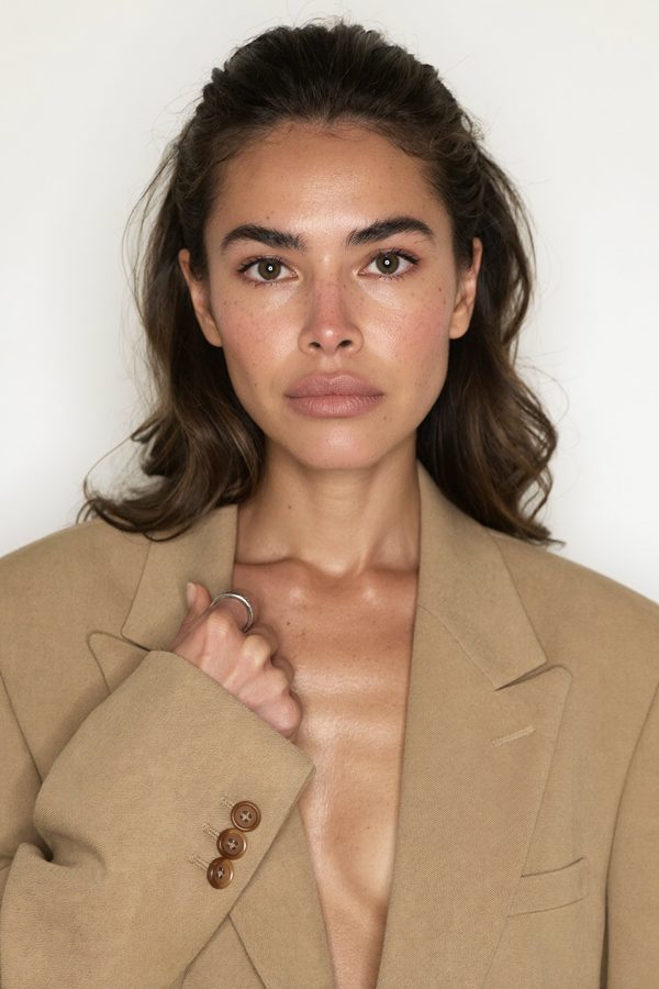 An AI brand model in an oversized camel blazer against a seamless white studio backdrop, looking directly into the lens