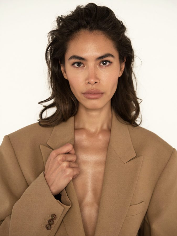 Variation: the AI brand model straight on to camera with her chin lifted, camel blazer, white backdrop