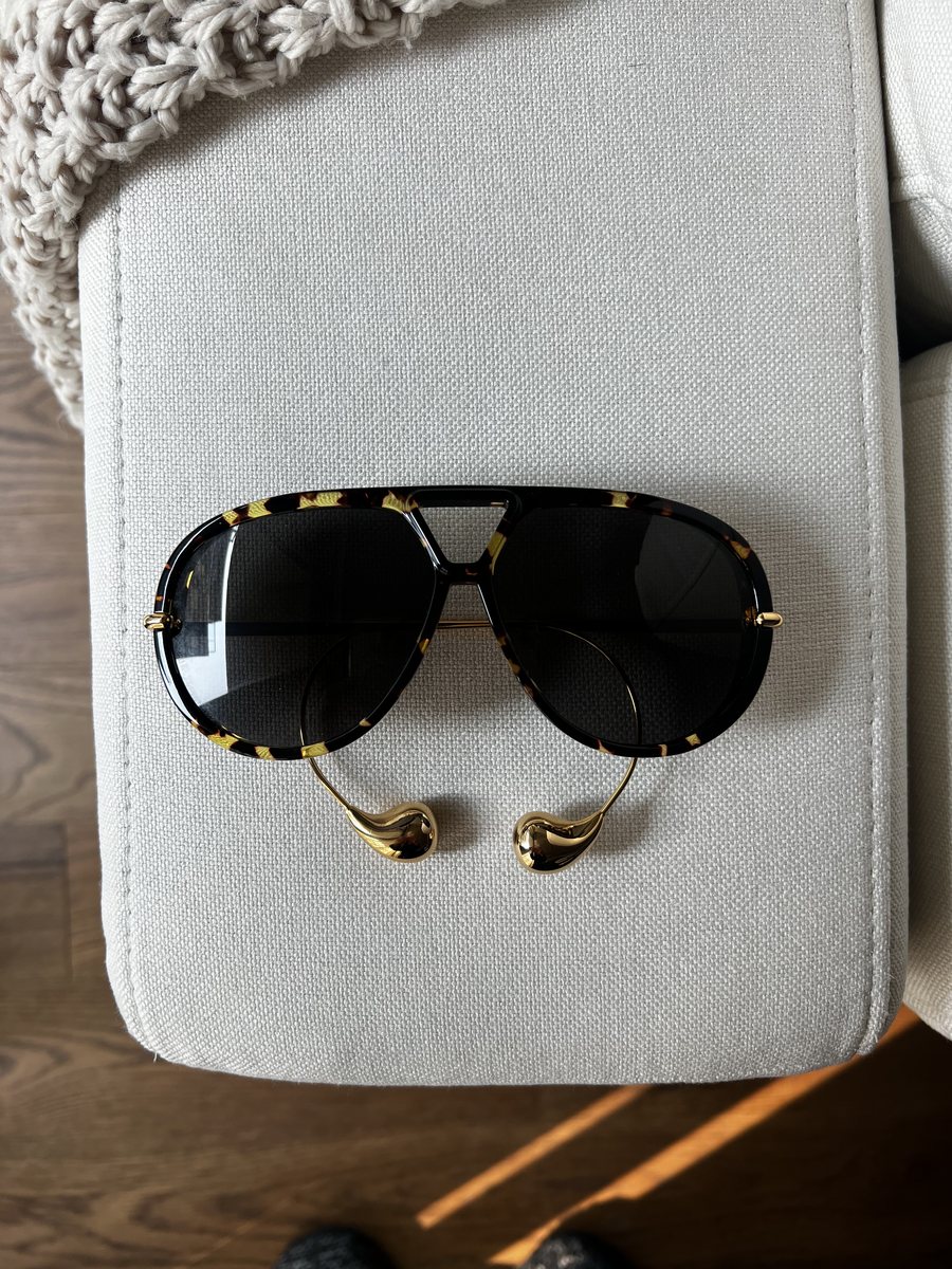 Tortoiseshell and gold sunglasses photographed on a linen chair arm with a phone, in daylight, with a knitted throw and a wood floor in frame