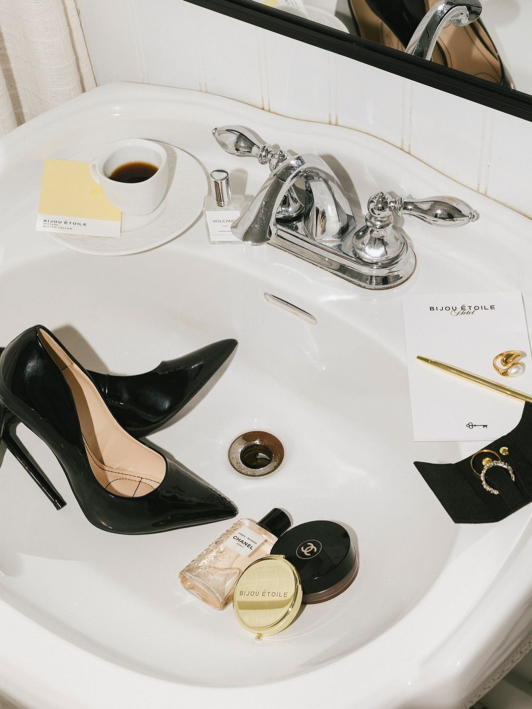 Black heels, cosmetics, jewelry and a coffee cup arranged around a white bathroom sink.