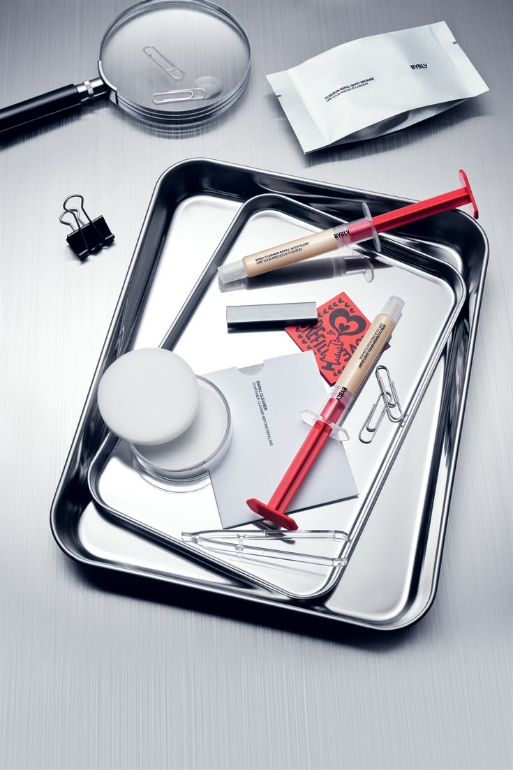 Beauty products and red syringe-shaped refill packaging arranged on reflective steel trays.