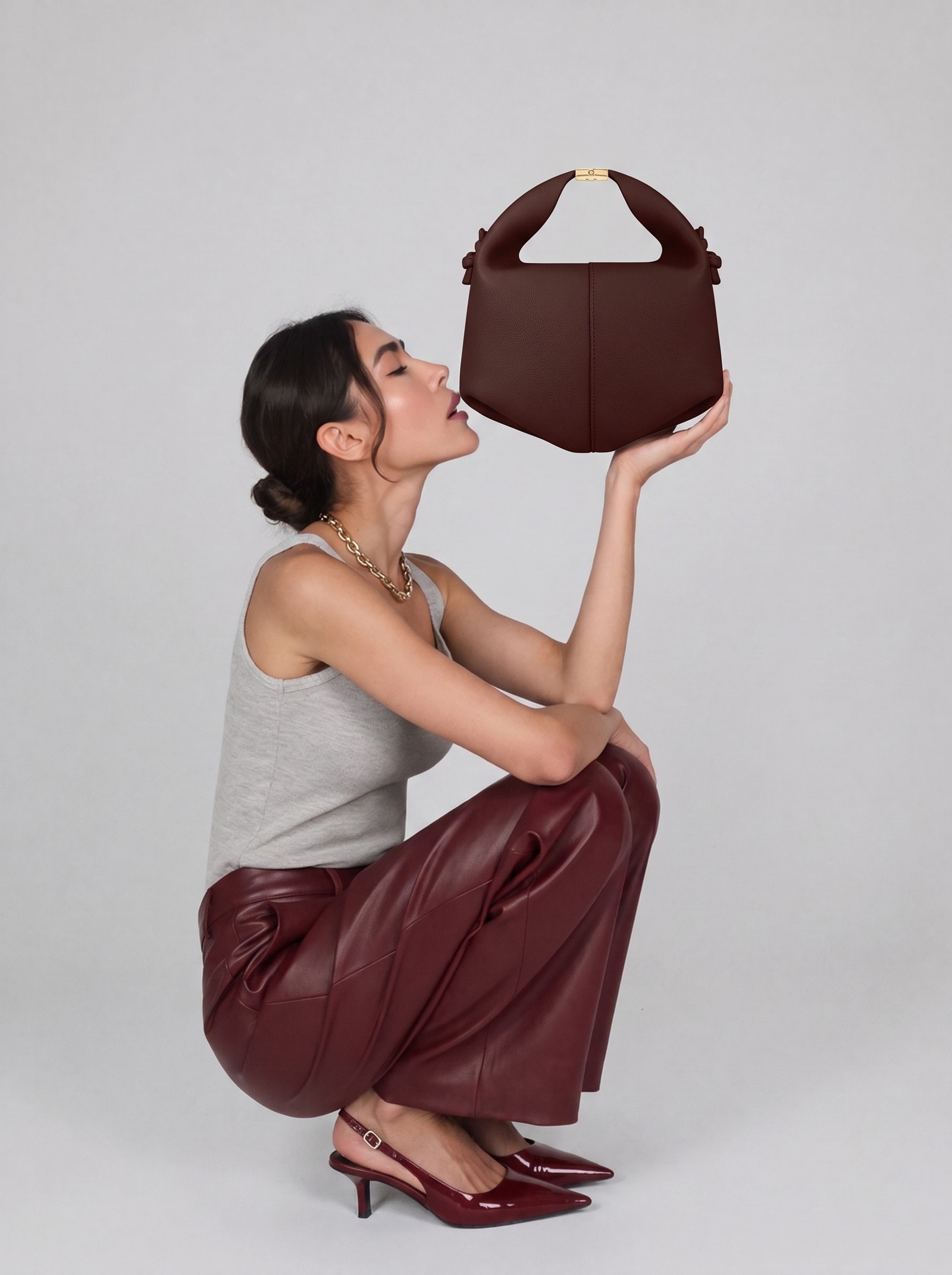 AI model wearing mustard gloves and holding a burgundy oxblood pebbled-leather top-handle bag, front-facing, studio editorial shot