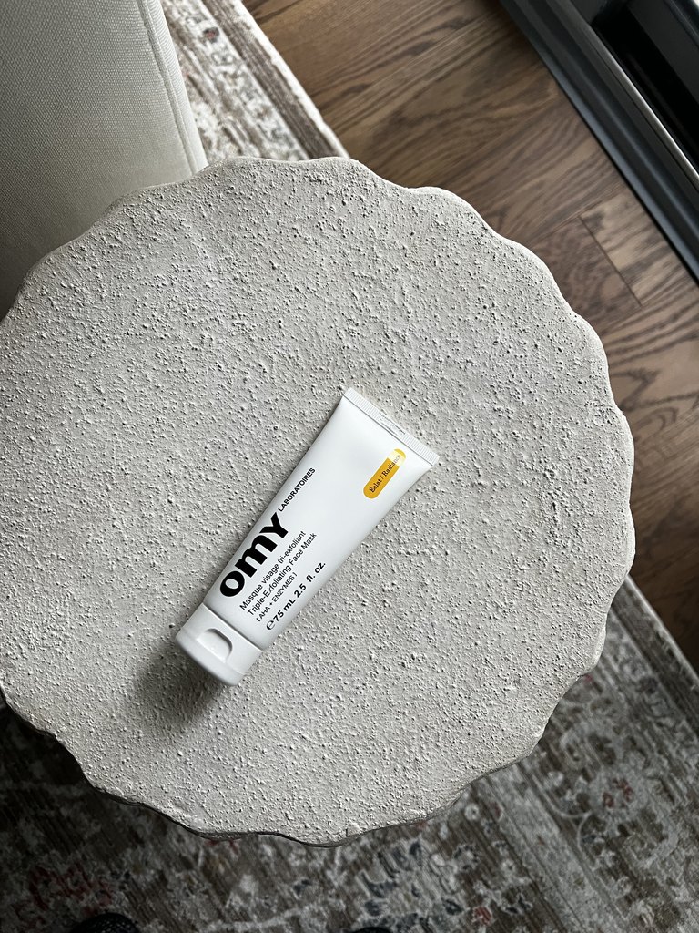 A white skincare tube lying on a textured cream plaster side table, photographed from above on a wooden floor