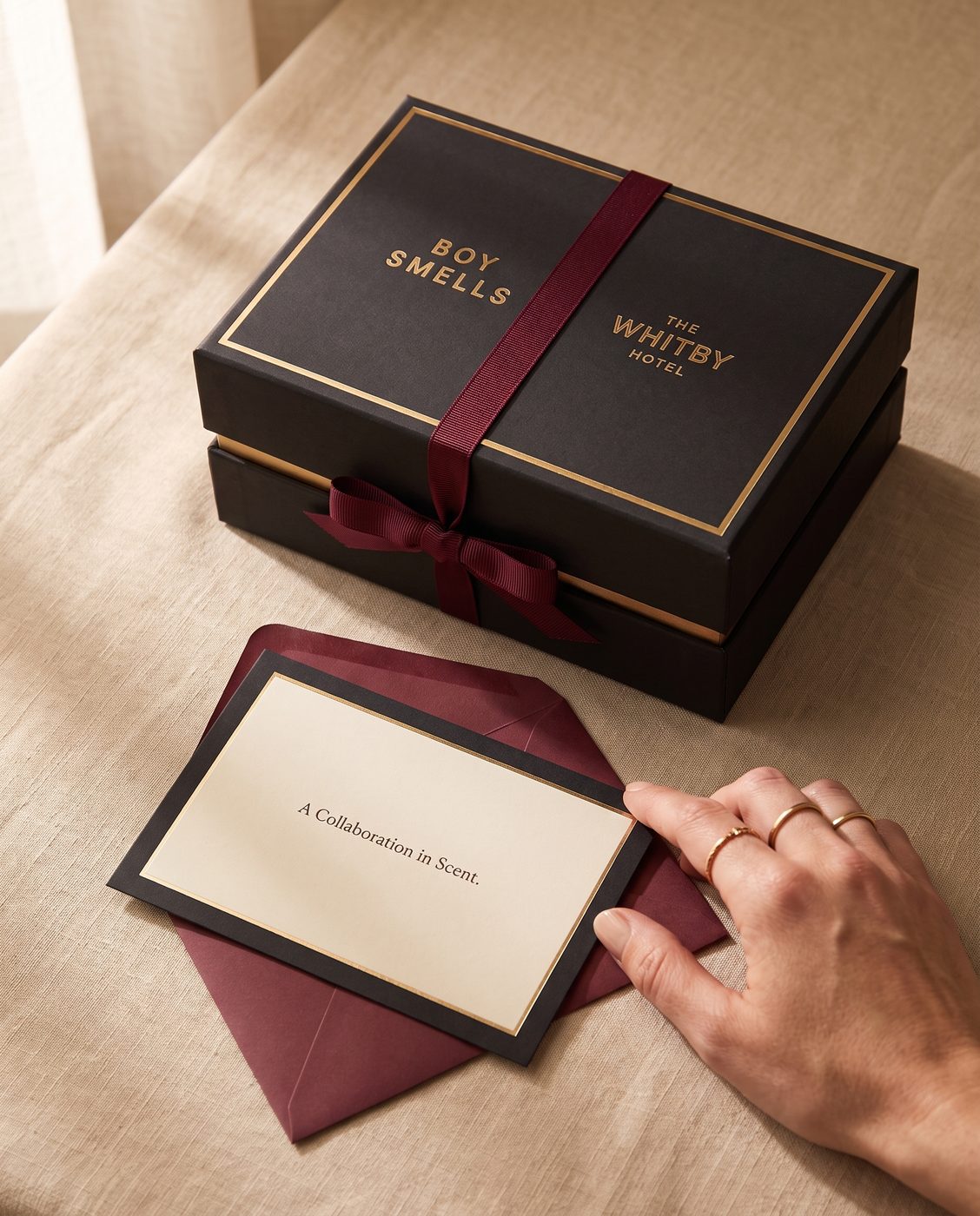 A co-branded gift box in black and gold with Boy Smells and The Whitby Hotel both stamped on the lid, tied with a burgundy ribbon, beside a card reading A Collaboration in Scent