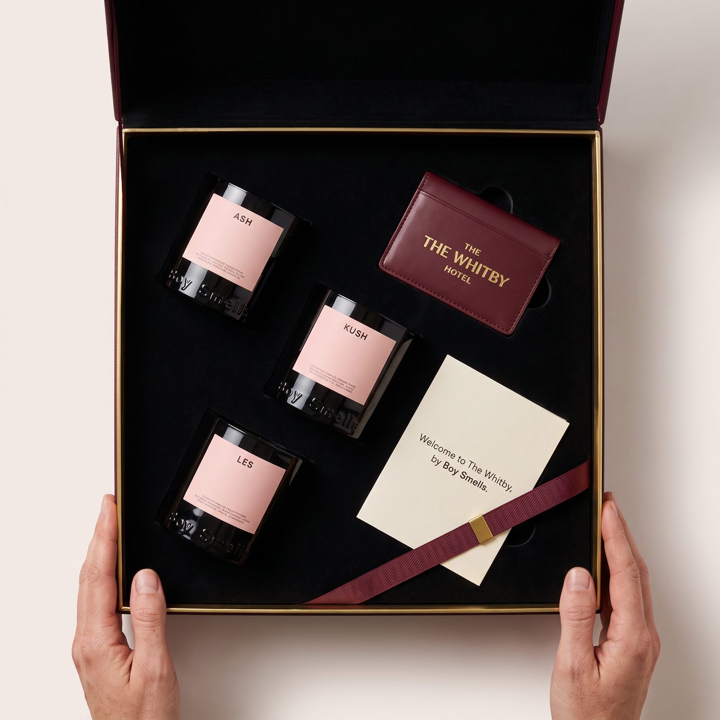 Overhead shot of the collaboration kit as it would ship: three Boy Smells candles, a Whitby Hotel card case, and a welcome card, styled inside an open black gift box