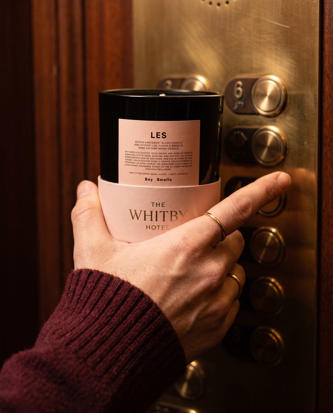 Second Instagram launch tile showing a hand in a burgundy sweater holding a Boy Smells Les candle in a pink Whitby Hotel sleeve, next to an elevator button panel